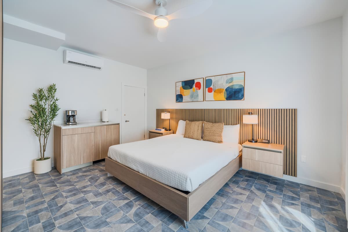 A modern hotel room with a bed, bedside tables, a potted plant, and abstract artwork on the wall.