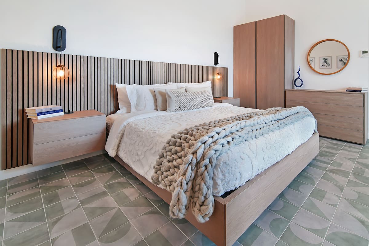A modern bedroom with a wooden headboard, a large bed with a chunky knit blanket, a bedside table with books, and a wooden wardrobe.