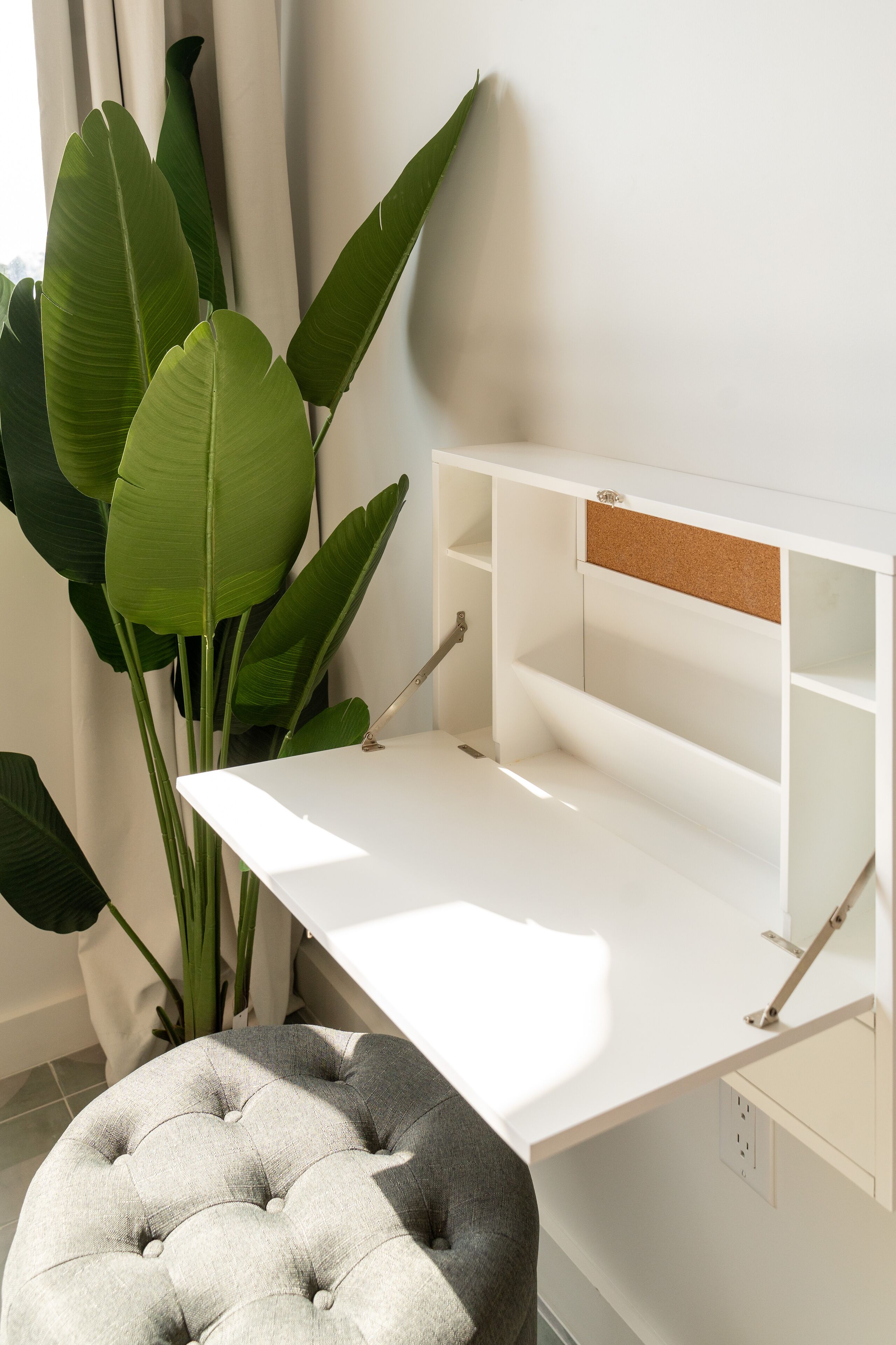A white desk with a corkboard, a grey cushioned stool, and large green leaves in the background.