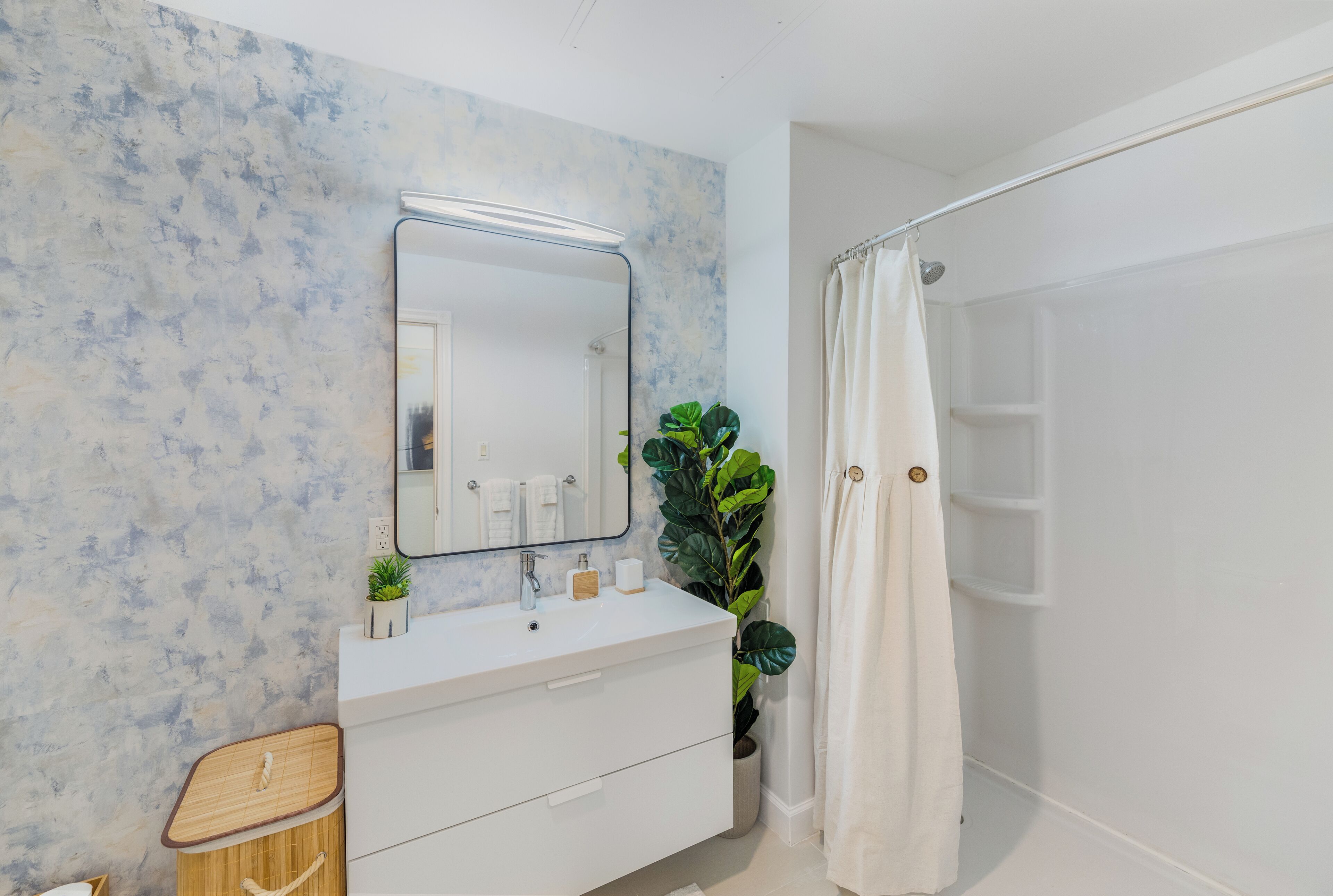 Et badeværelse med en marmorvæg, et hvidt toiletbord, et spejl, en bruser med gardin og en potteplante.