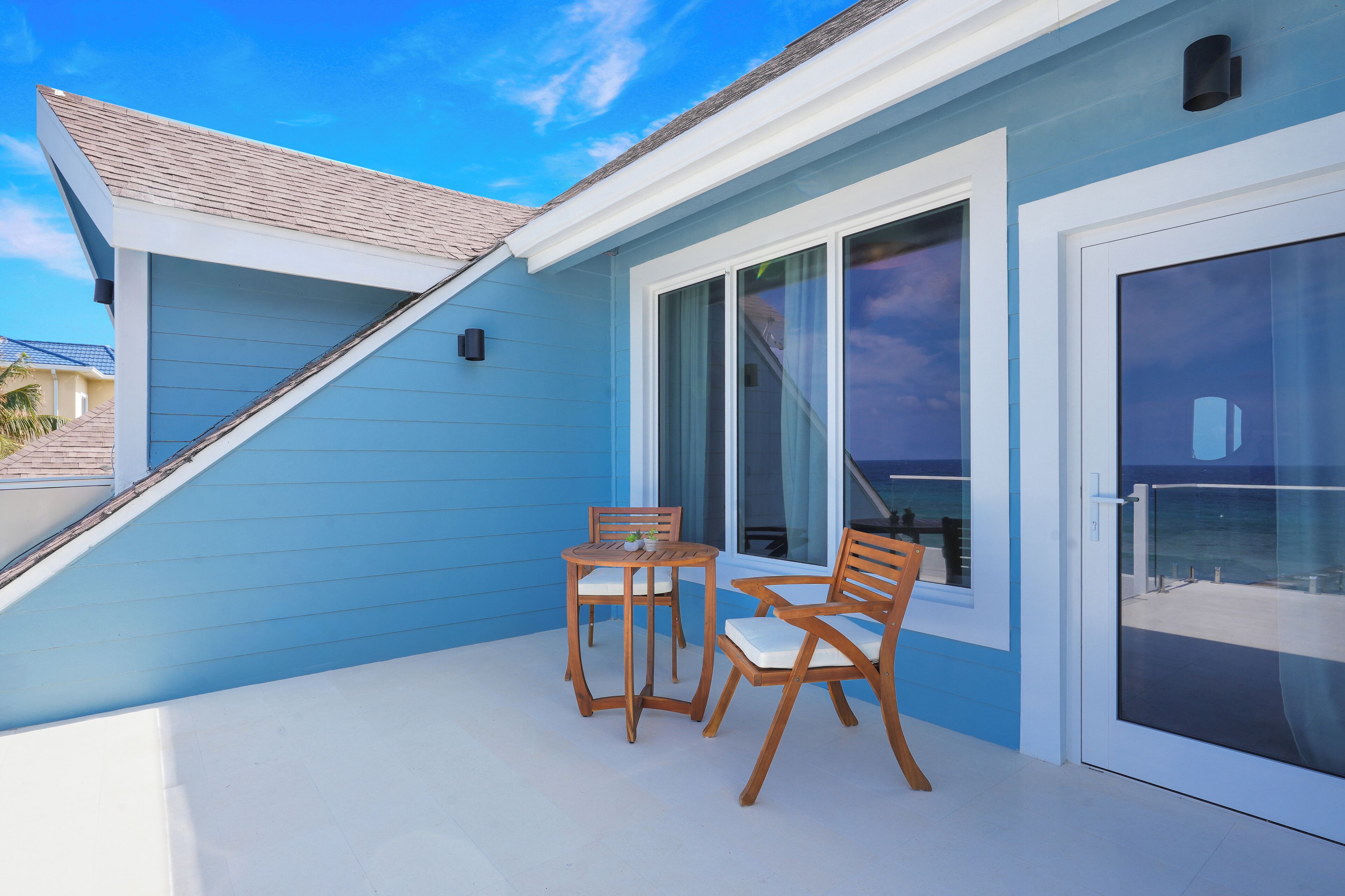 A blue building with a covered patio area featuring two wooden tables and chairs, large windows, and a view of the ocean.