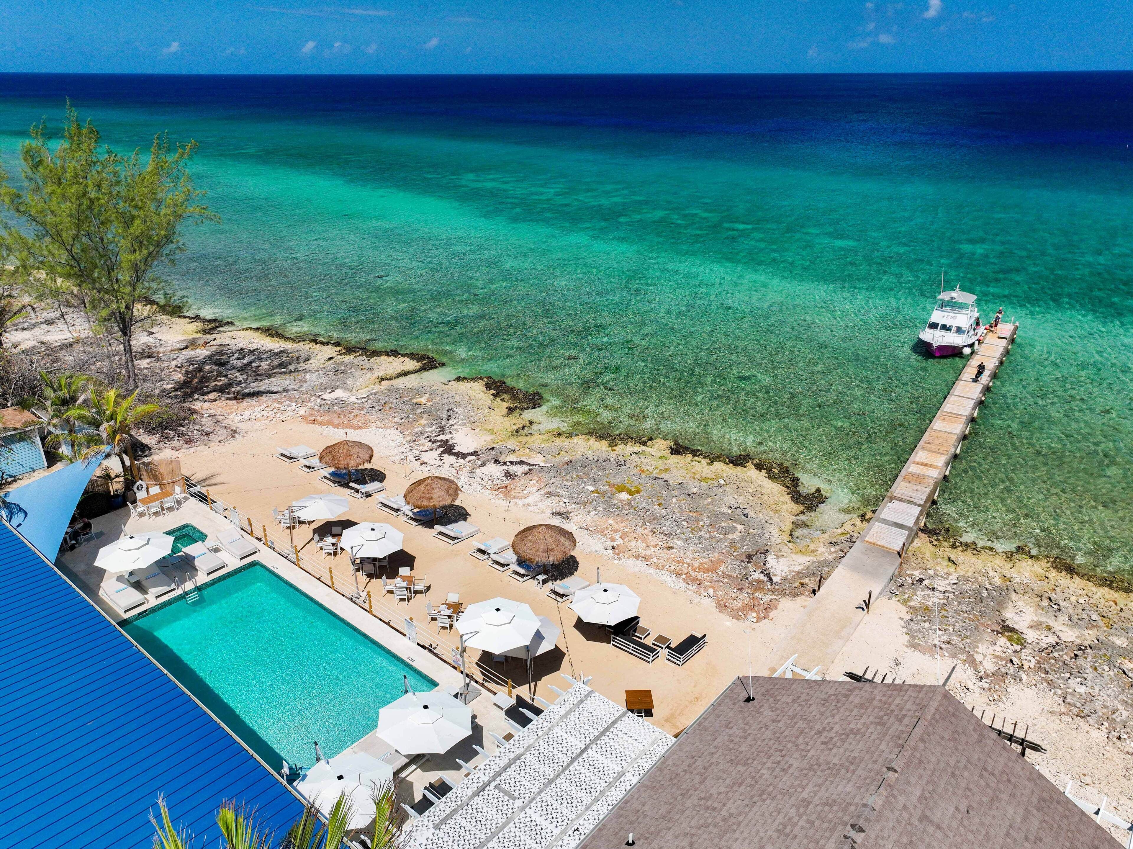 Un resort frente a la playa con piscina, tumbonas y un muelle que se adentra en el mar.