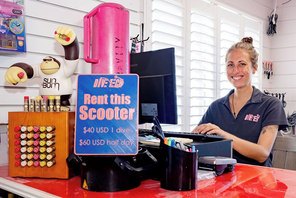 A person standing behind a counter with a sign offering scooter rentals for $40 USD for one dive and $60 USD for half a day.
