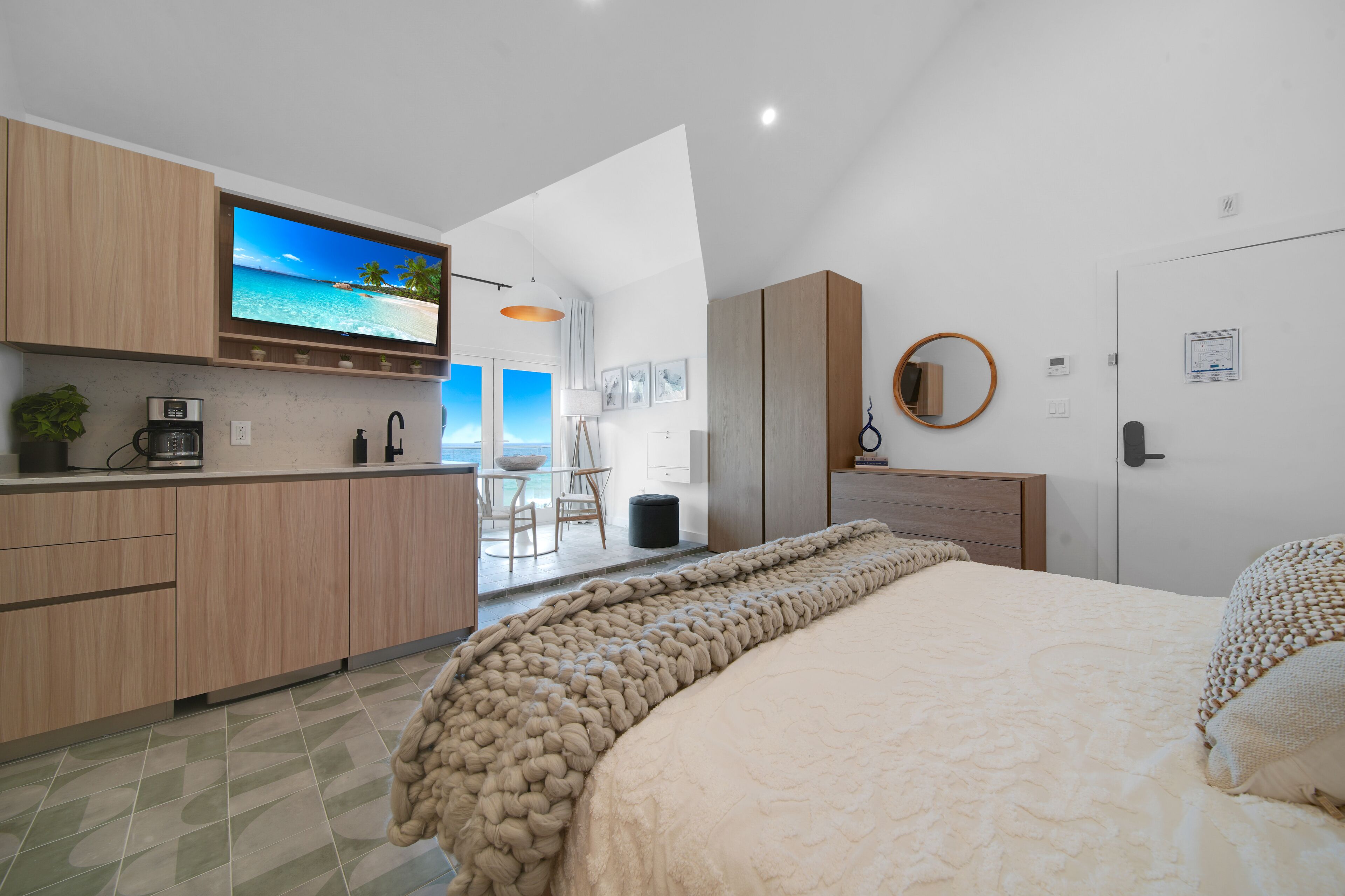 Ein modernes Schlafzimmer mit einem großen Bett, einem an der Wand montierten Fernseher und Blick auf den Strand durch das Fenster.