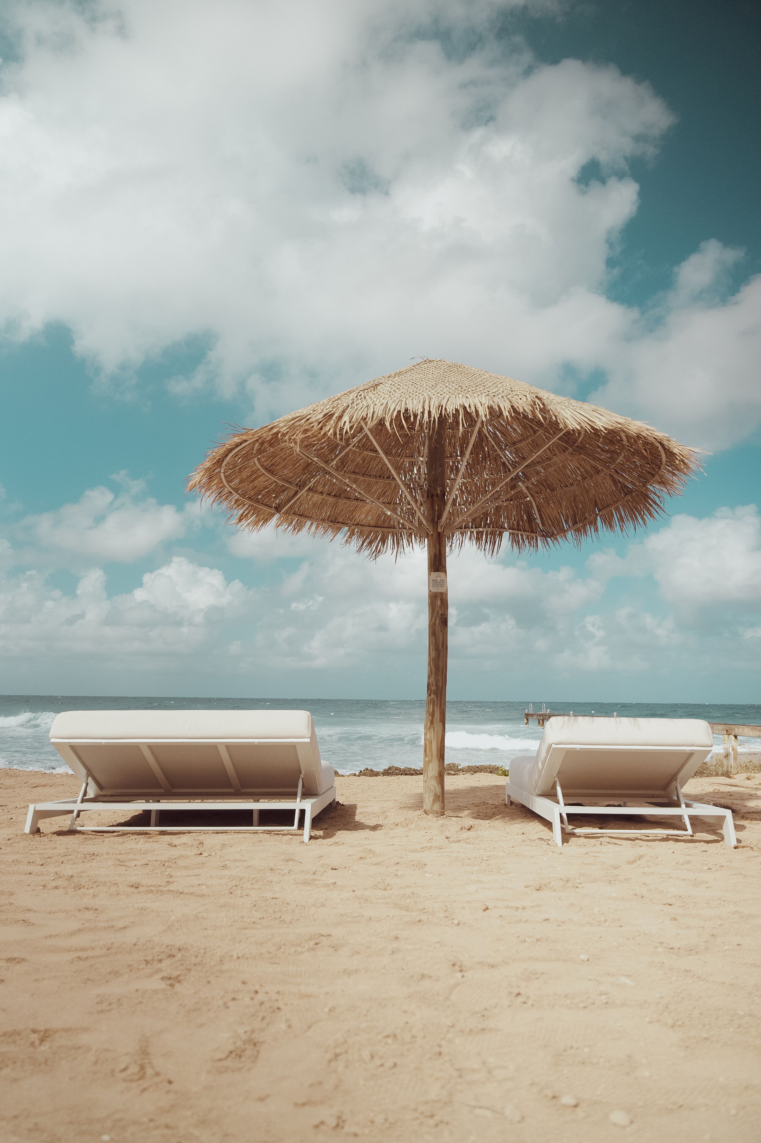 Two beach chairs under a straw umbrella on a sandy beach with the ocean in the background.