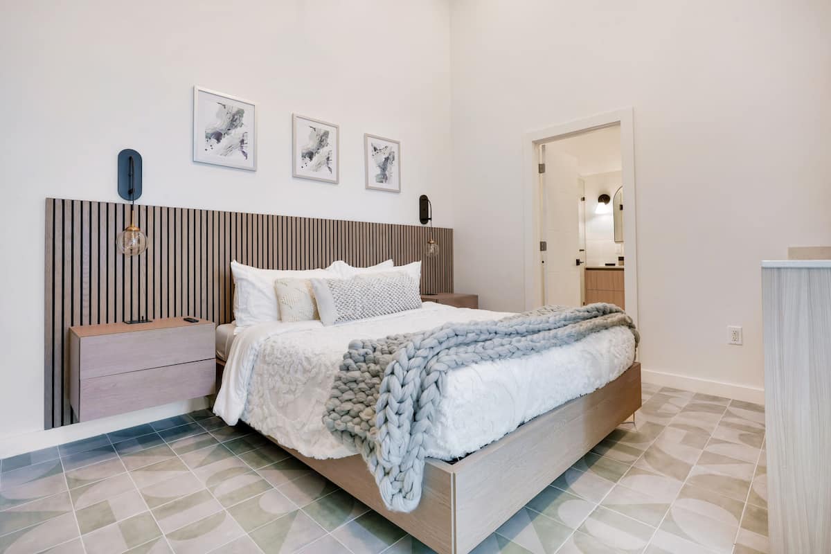 A modern bedroom with a wooden headboard, a large bed with a grey blanket, a nightstand, and a wall-mounted lamp.