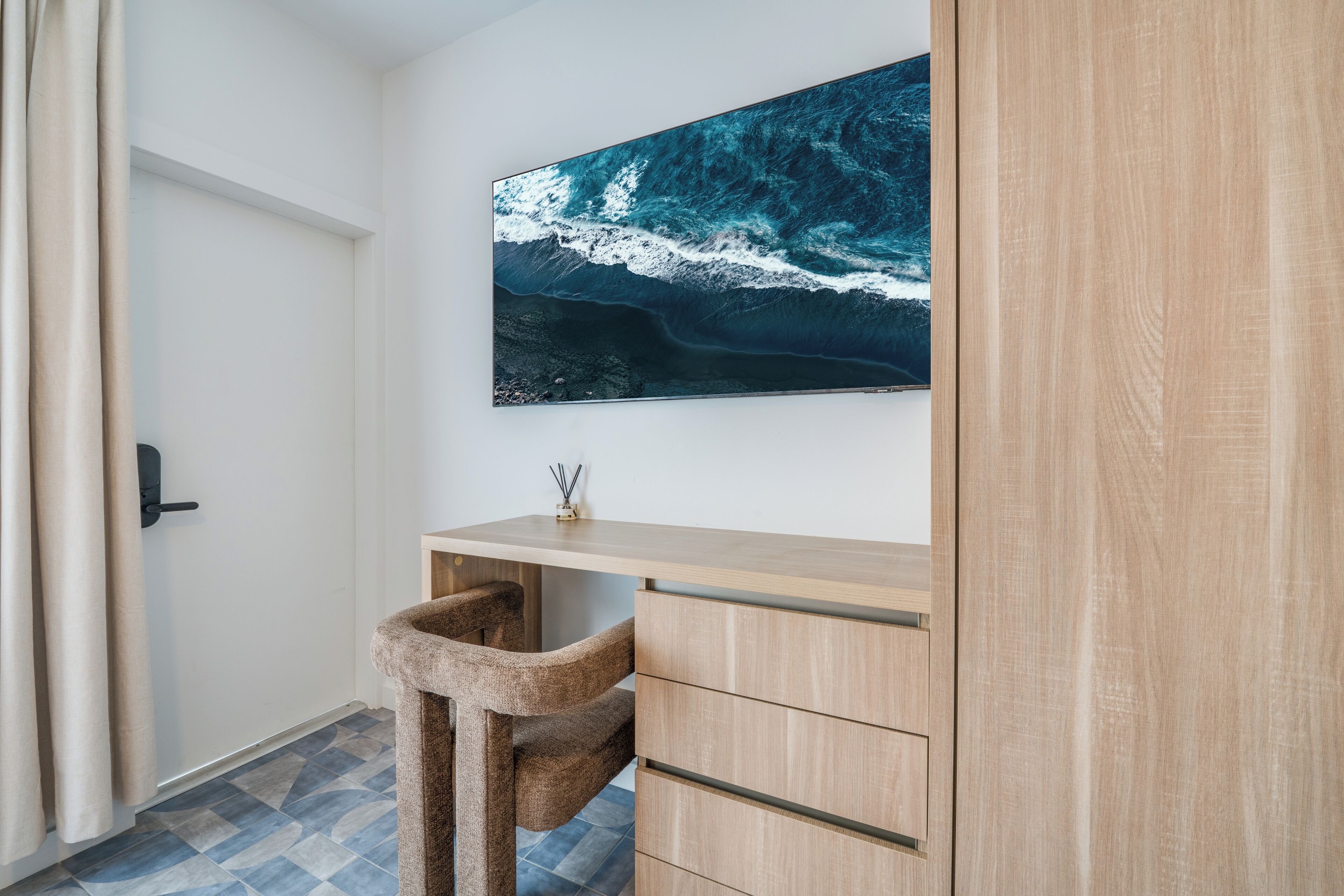 Ein Zimmer mit Schreibtisch aus Holz, einem Stuhl, einem großen gerahmten Bild einer Welle und einer Tür mit Vorhängen.