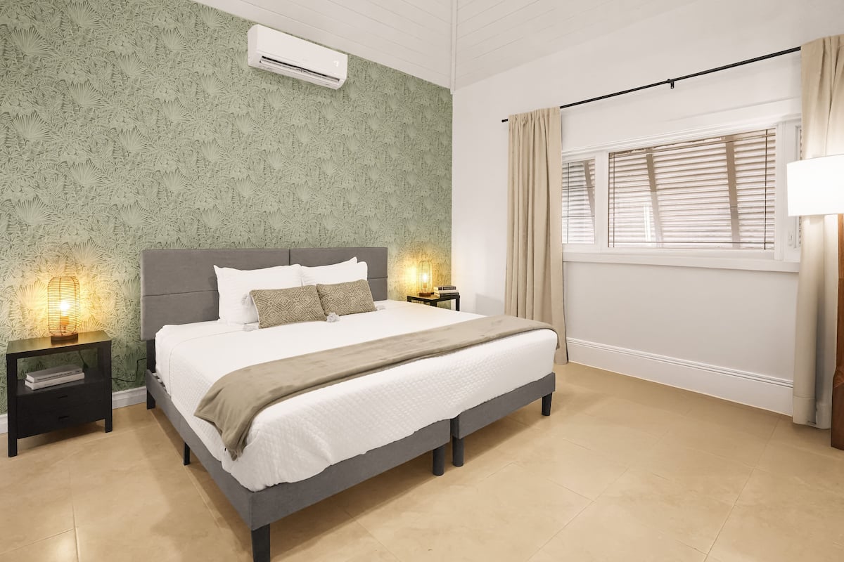 A modern bedroom with a large bed, two bedside tables with lamps, a window with blinds, and a patterned wallpaper.