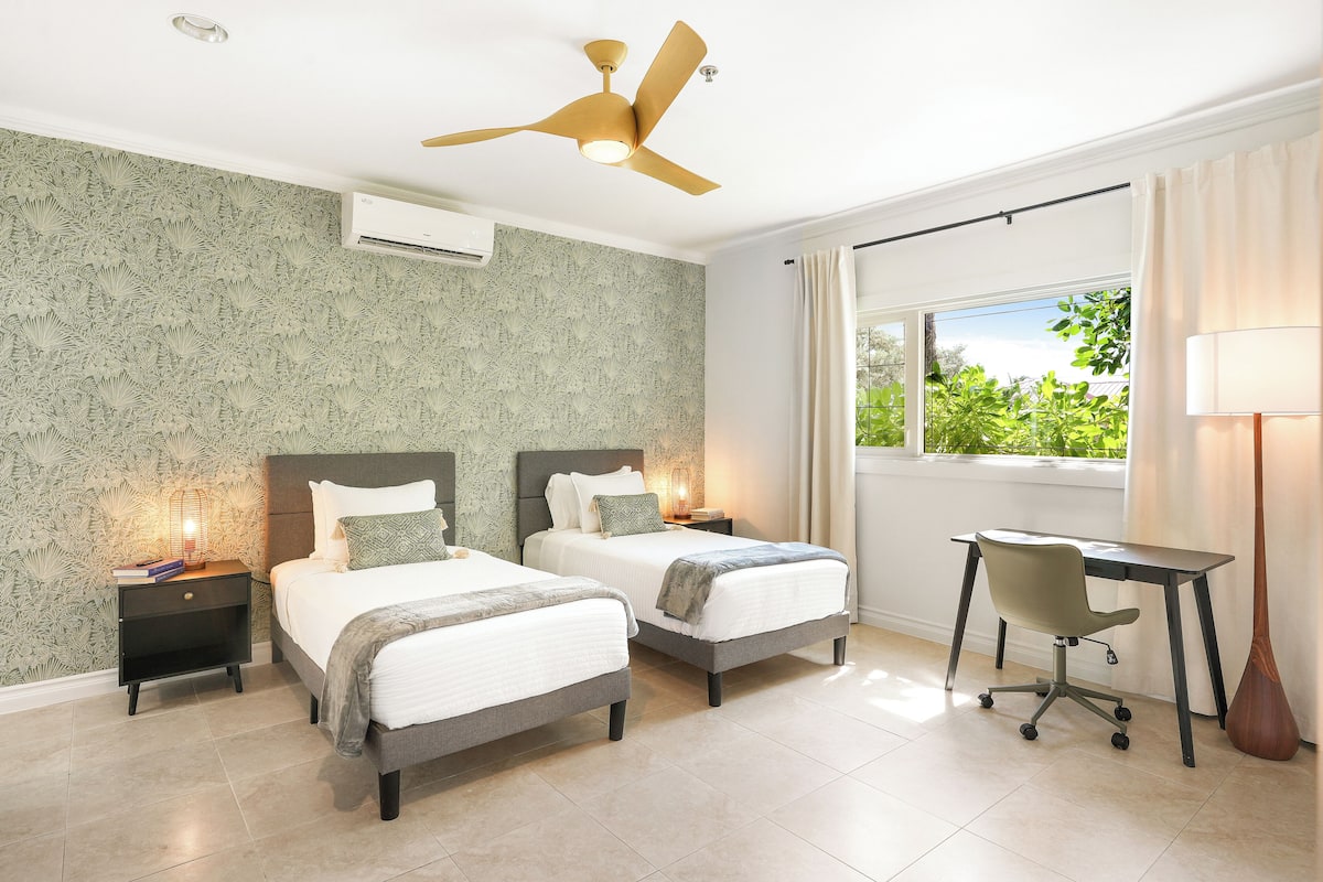 A bedroom with two beds, a desk, a chair, a ceiling fan, and a window with a view of greenery.