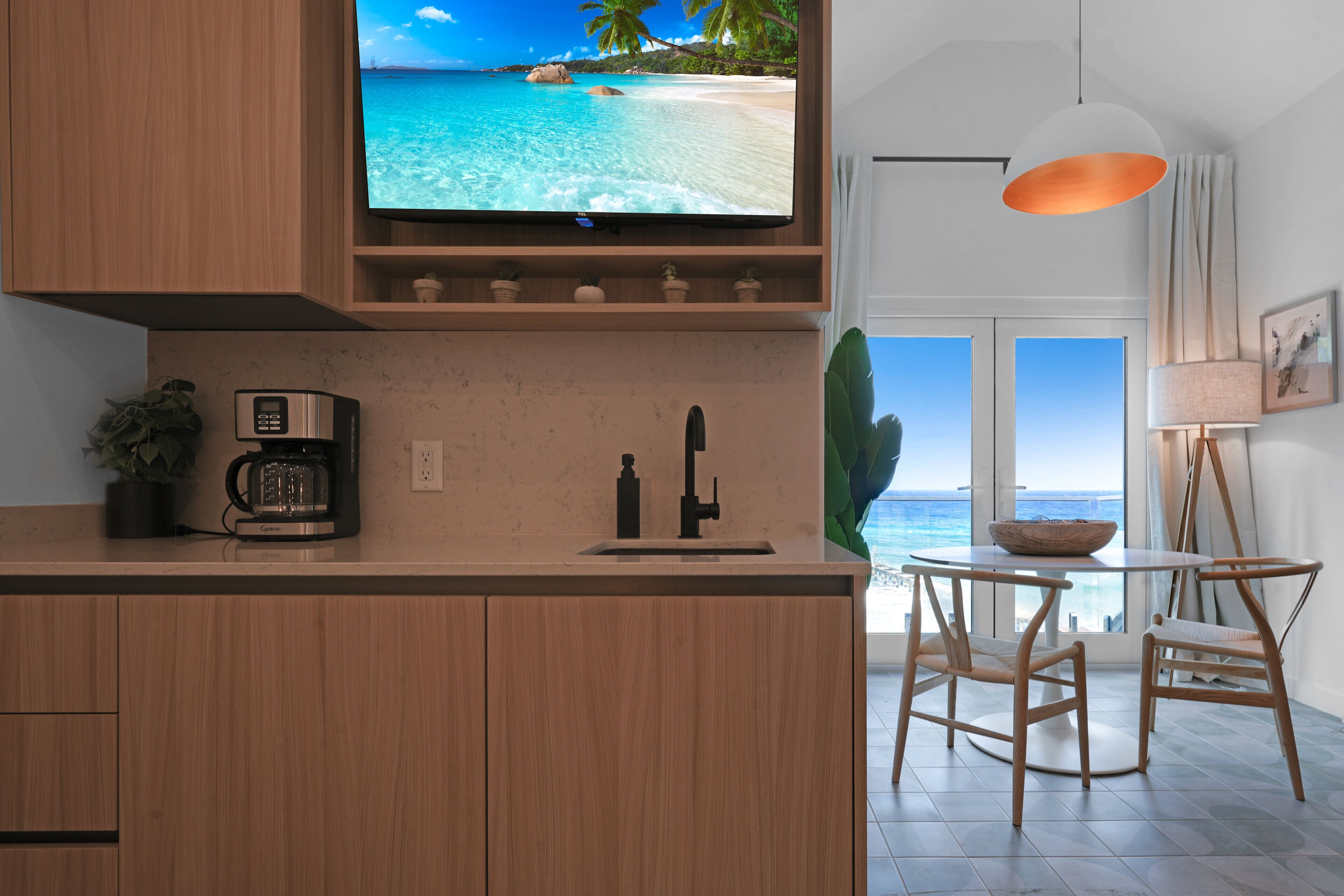 Eine moderne Küche mit Holzausstattung, einer Kaffeemaschine und einem Fernseher, der eine Strandansicht zeigt. Es gibt einen Essbereich mit Tisch und Stühlen sowie einen Blick auf den Ozean durch große Fenster.