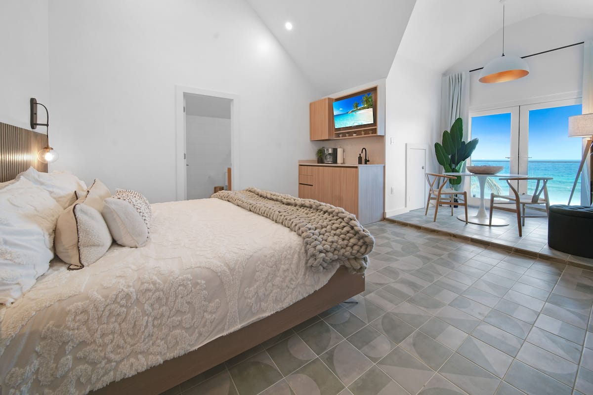 A bedroom with a large bed, a TV, a kitchenette, and a balcony with a view of the sea.