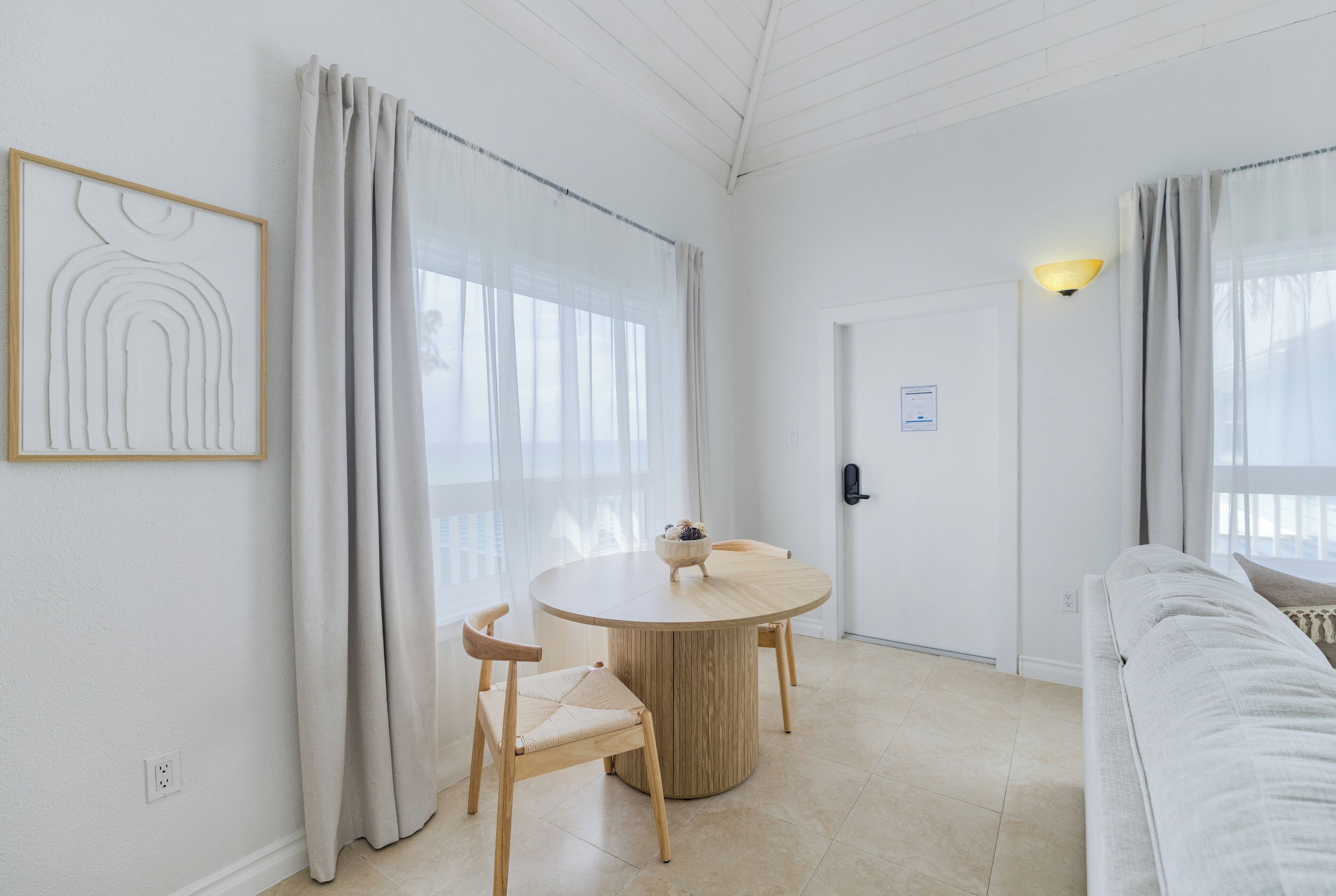 Una habitación moderna y minimalista con una mesa redonda de madera, dos sillas de madera, un sofá blanco y una obra de arte enmarcada en la pared.