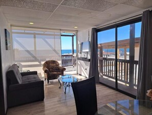 Ein Zimmer mit einer Couch, einem Sessel und einem kleinen Tisch. Ein Balkon mit Geländer und ein an der Wand befestigter Fernseher sind vorhanden.
