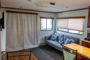 A compact RV interior with a sofa, a small table, a window with blinds, and a television mounted on the wall.