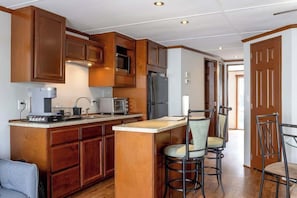 A kitchen with wooden cabinets, a refrigerator, a microwave, and a coffee maker. There are two bar stools and a dining table with chairs.