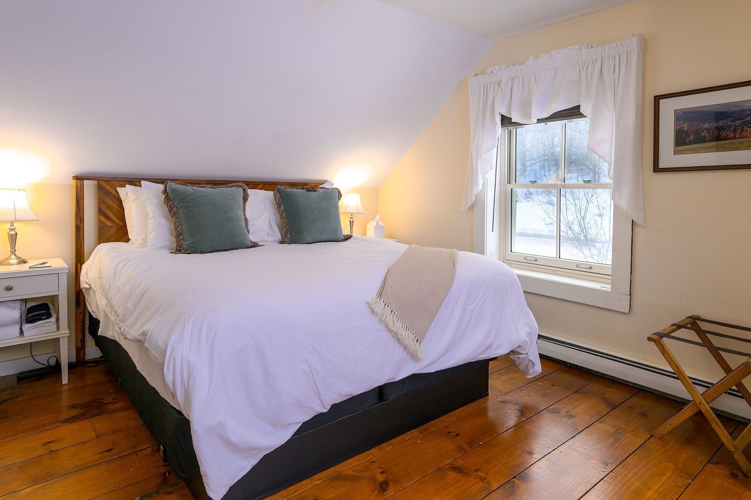 A neatly made bed with white linens and two pillows, a bedside table with a lamp, and a window with white curtains.