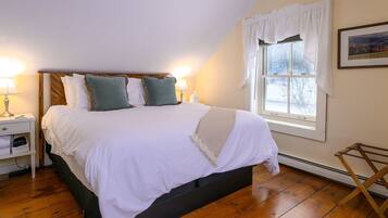 A neatly made bed with white linens and two pillows, a bedside table with a lamp, and a window with white curtains.