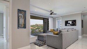 A modern living room with a grey sofa, a ceiling fan, and a large window offering a view of the cityscape.