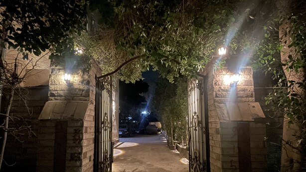 Une entrée nocturne, avec des murs en pierre et des portails en métal, qui mène à un sentier faiblement éclairé.