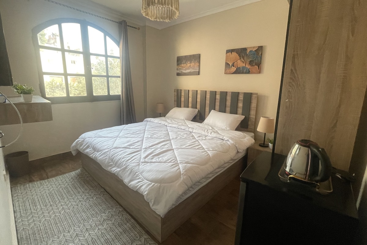 A neatly arranged bedroom with a bed, bedside lamps, a coffee maker, and artwork on the wall.
