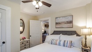 Een slaapkamer met een bed, een plafondventilator, een nachtkastje, een lamp, een dressoir, een spiegel en een schilderij aan de muur.