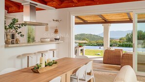 A modern kitchen with a wooden table and bar stools, a large window with a view of the outdoors, and a dining area with wicker furniture.