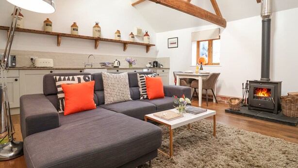 A cozy living room with a gray sectional sofa, a wooden ceiling, a fireplace, and a kitchen in the background. - The Nook (Halesworth)
