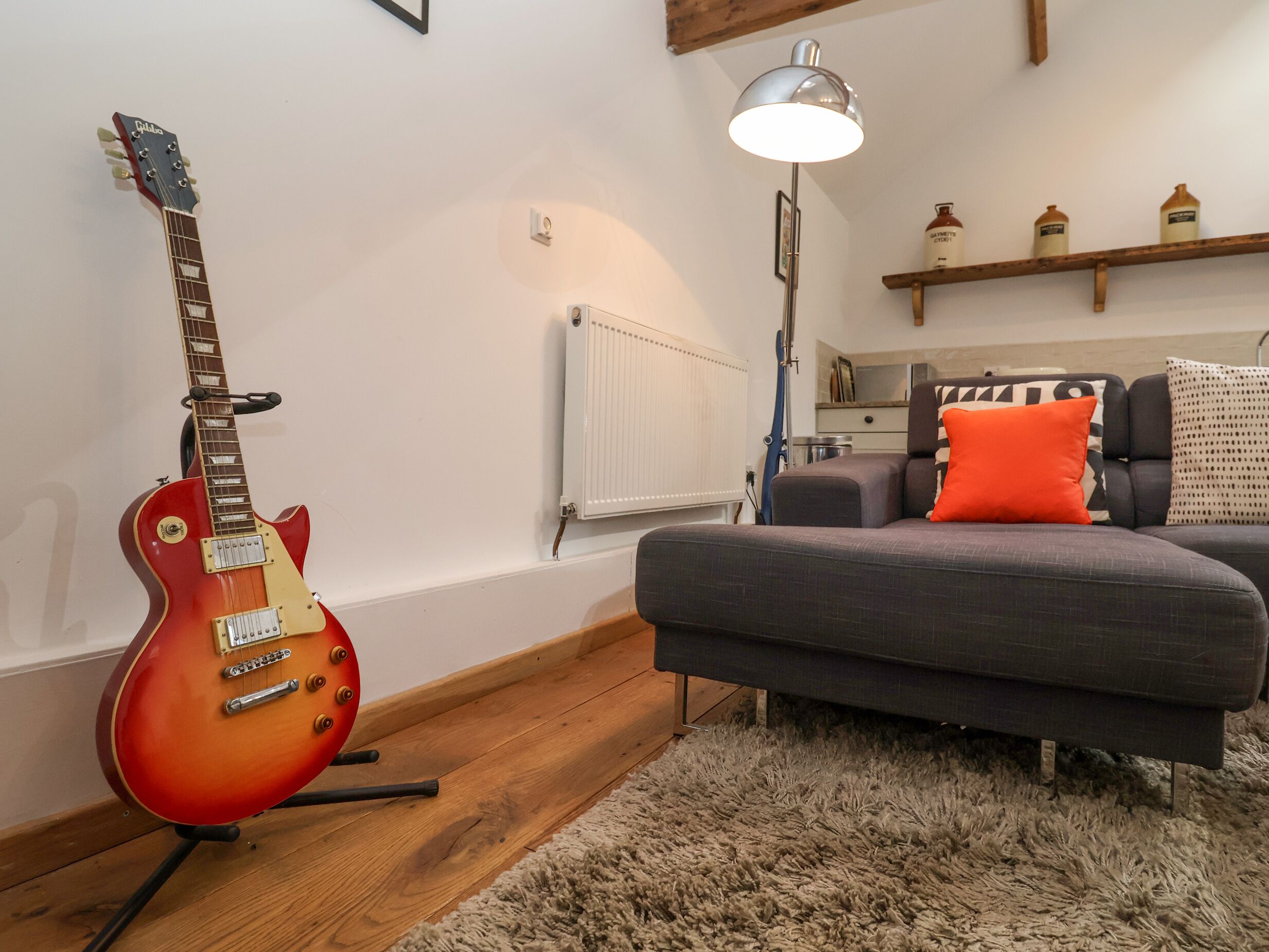 Una sala de estar con una guitarra eléctrica roja, un sofá gris con cojines naranjas, una alfombra mullida, piso de madera, un radiador blanco y una estantería con frascos.