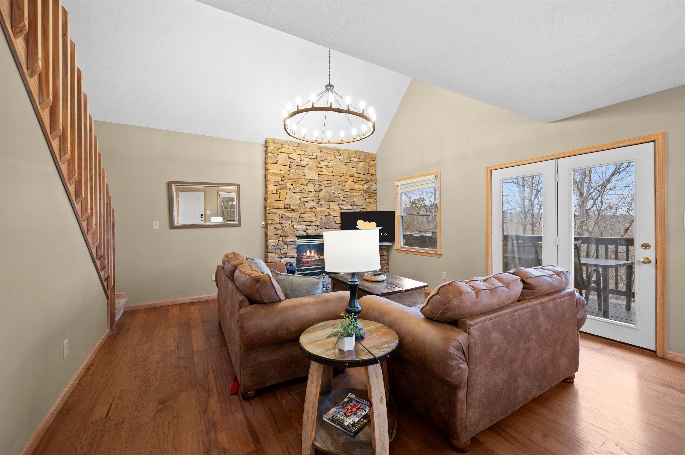 A cozy living room with a stone fireplace, leather sofas, a chandelier, and a wooden staircase.