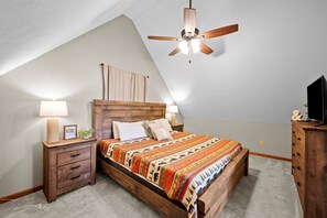A bedroom with a sloped ceiling, a wooden bed frame, a ceiling fan, and a nightstand with a lamp. - Cozy Hot Tub Chalet | Minutes to The Island (Pigeon Forge)