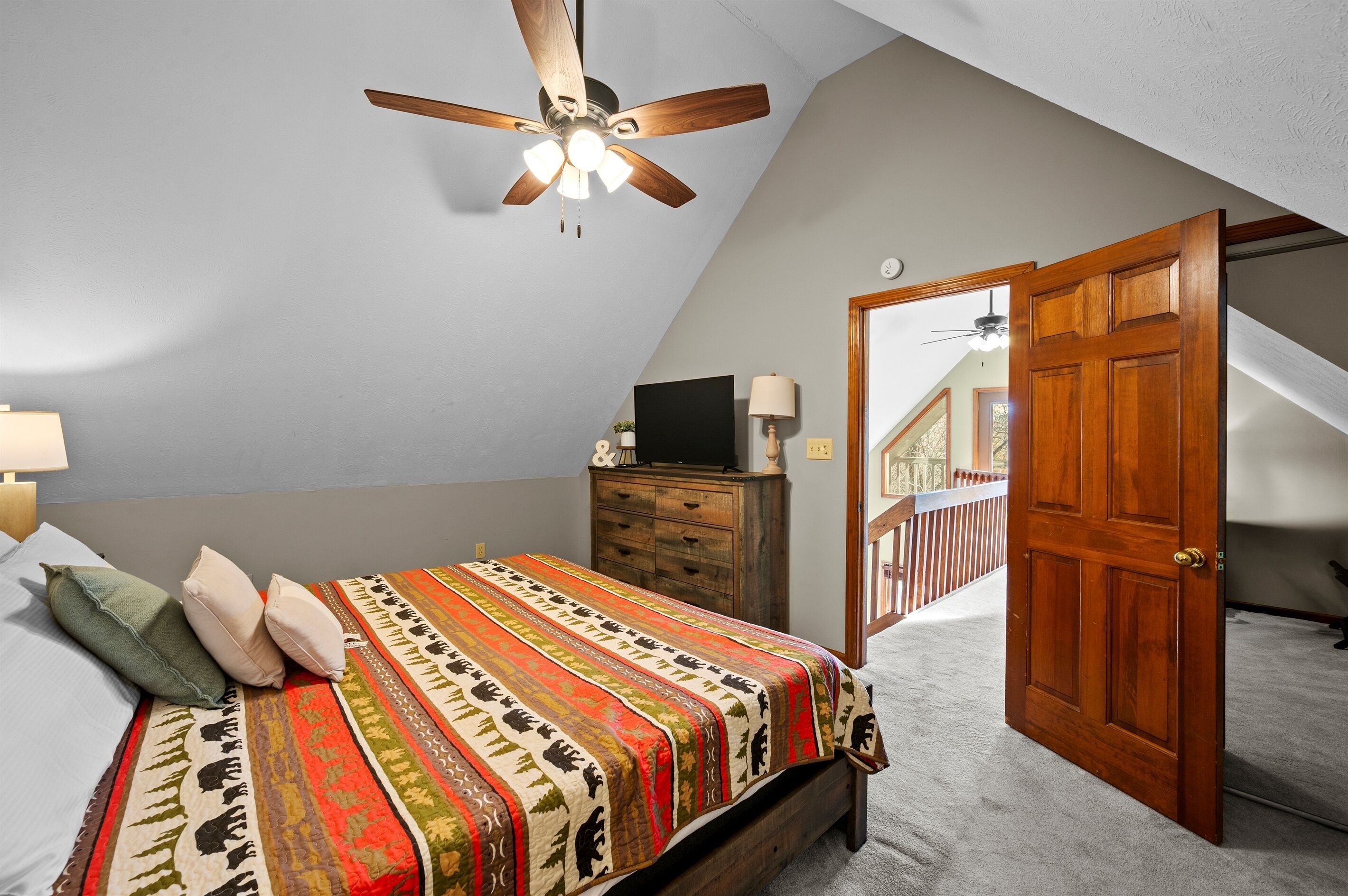Ein Schlafzimmer mit schrägem Dach, einem Bett mit gemustertem Überwurf, einer Holzkommode, einem Fernseher und einer Tür, die auf einen Balkon im Freien führt.