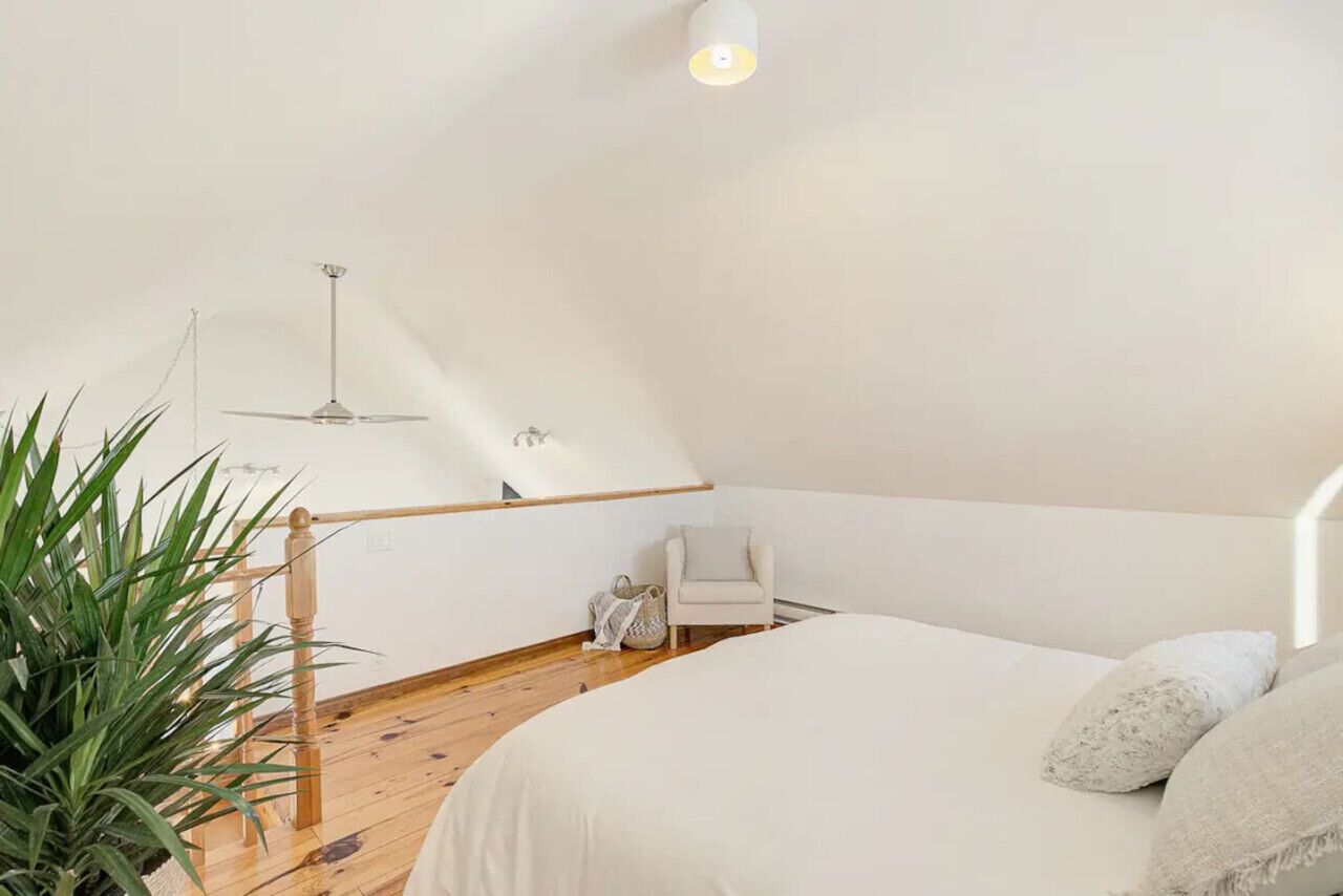 A bedroom with a bed, a chair, a hanging light fixture, and a wooden staircase.