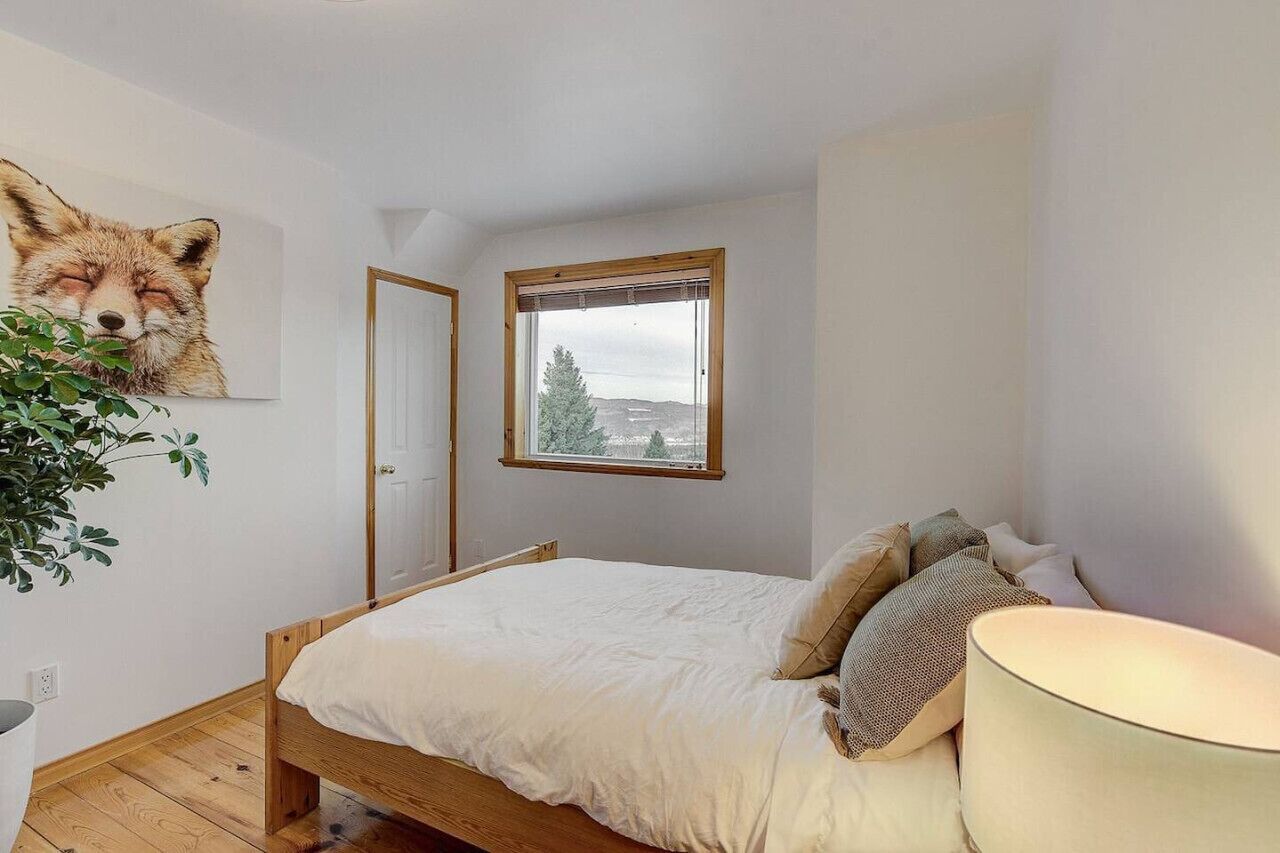 A bedroom with a bed, a window with a view, a door, and a wall-mounted fox painting.