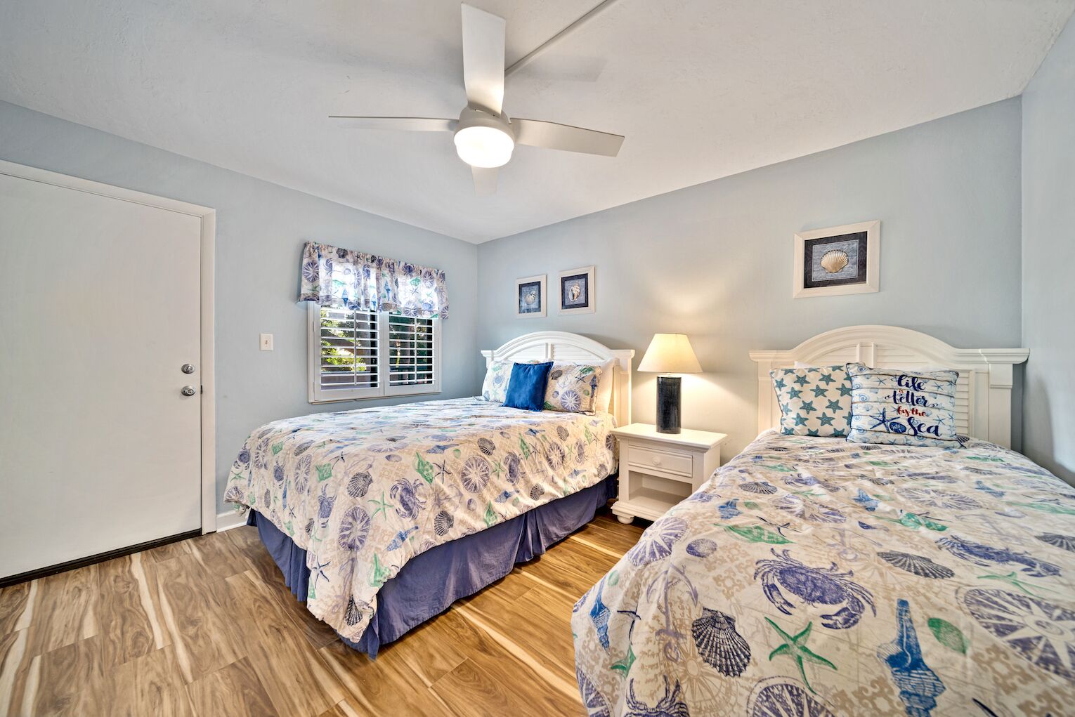 A bedroom with two beds, a ceiling fan, a nightstand with a lamp, and a window with shutters.