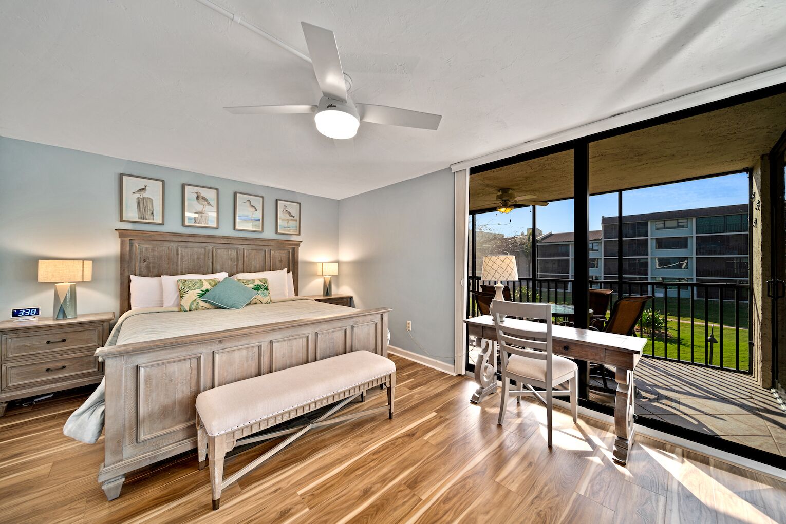 A bedroom with a bed, a nightstand, a desk, a chair, a ceiling fan, and a large window with a view of buildings.