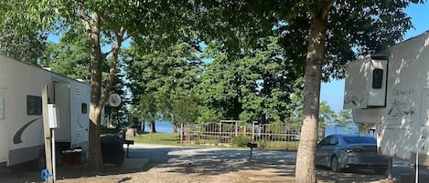 A gravel parking area with RVs and a car, surrounded by trees and a fence.