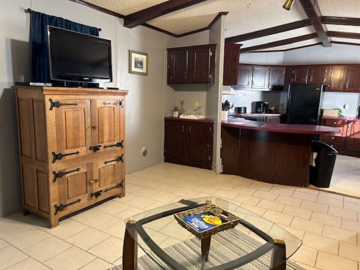A compact living space with a kitchenette, a wooden cabinet with drawers, a television on top, and a glass table with a magazine.