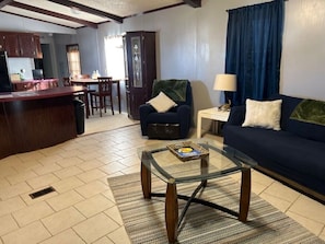 A living room with a glass coffee table, a sofa, a chair, and a lamp. There is a kitchen area in the background.