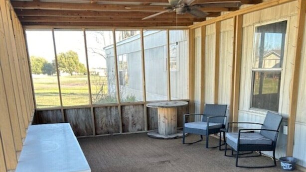 En overdækket terrasse med et bord og stole, en loftsvifte og udsigt til et hus gennem store vinduer.