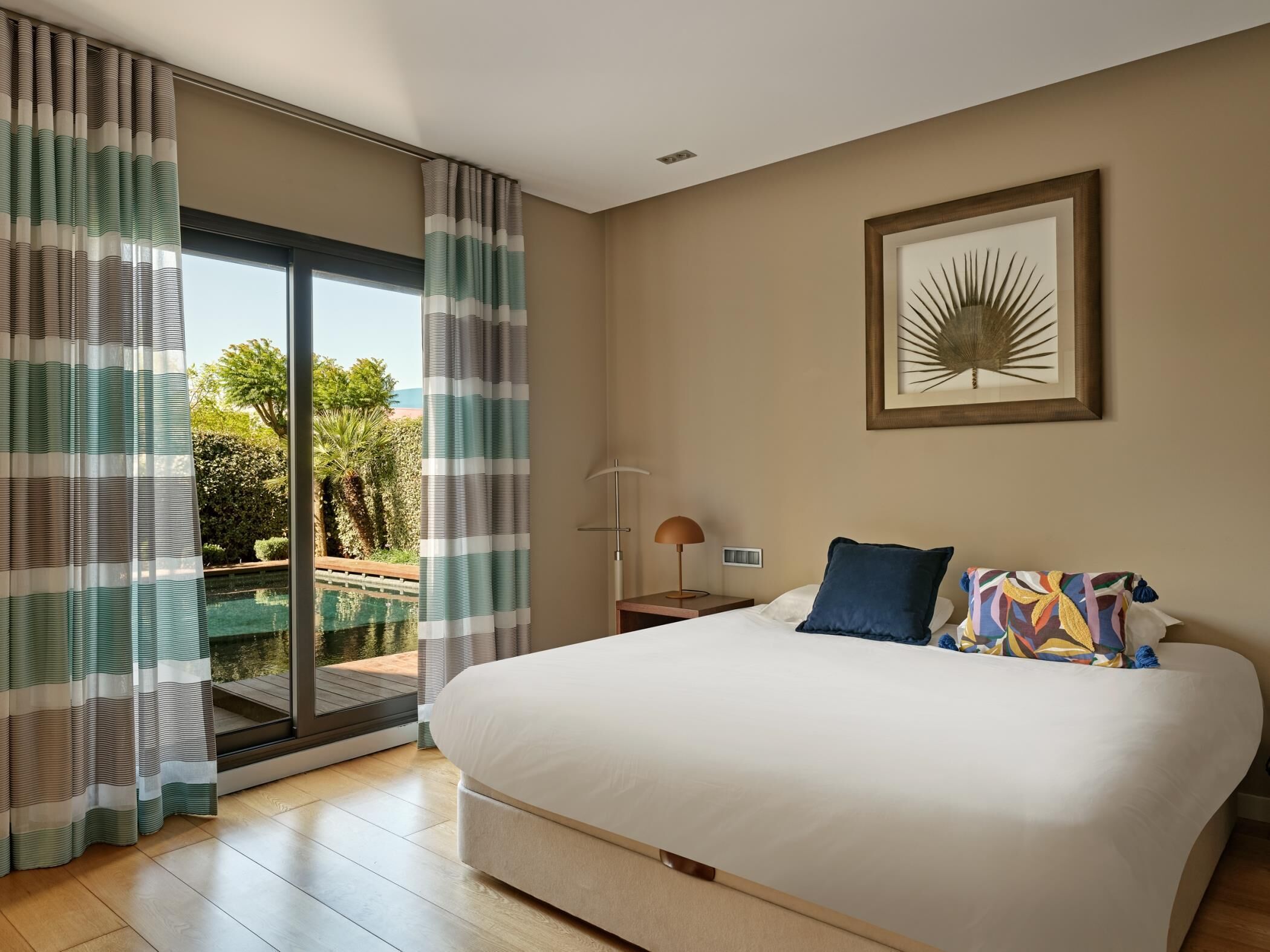 Ein Schlafzimmer mit einem großen Bett, einem Nachttisch, einer Lampe, einem gerahmten Bild an der Wand und Blick auf einen Pool durch das Fenster.