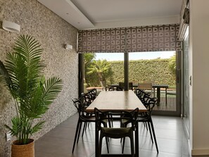 A dining area with a long table, black chairs, a large potted plant, and a patterned window blind.
