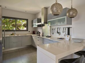 Een moderne keuken met witte kasten, een groot raam, hangende keukengerei en twee sierlijke lampen.