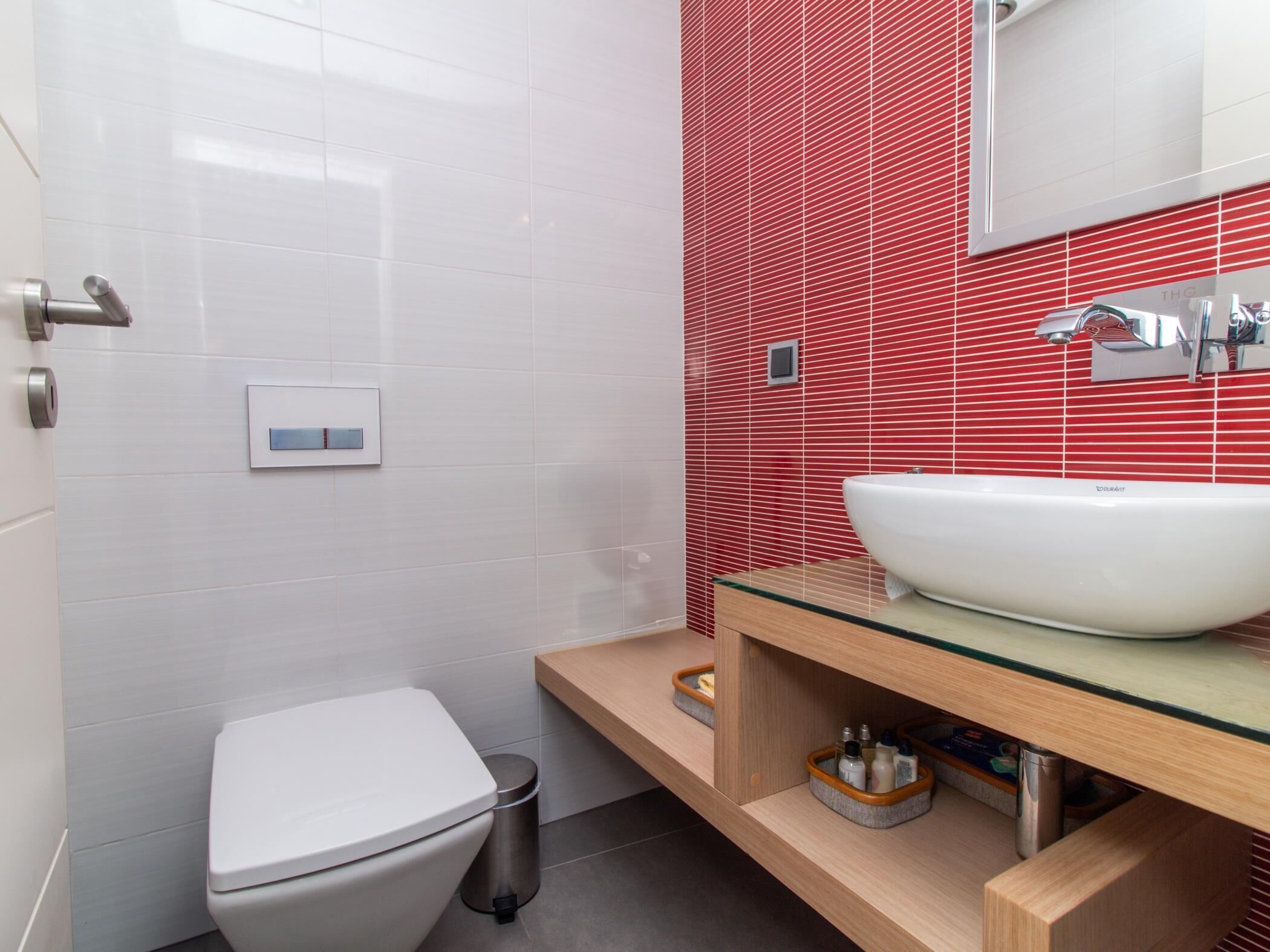 Un bany modern amb un vàter blanc, un lavabo de vidre i una paret de rajoles vermelles.