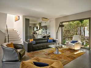Een moderne woonkamer met een bank, fauteuil en salontafel, een trap en een keuken op de achtergrond.