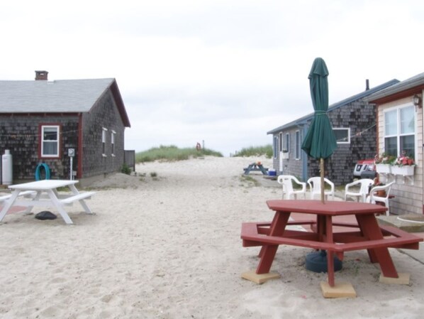 A sandy beach area with picnic tables and umbrellas, surrounded by stone buildings with windows and doors.
