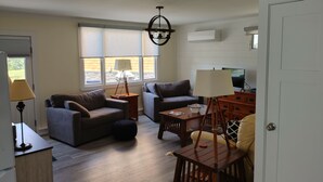 A living room with a sofa, armchair, wooden coffee table, and a lamp. There is a window with blinds, a ceiling fan, and a door.