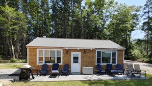 A small wooden cabin with a porch, several Adirondack chairs, and a grill.