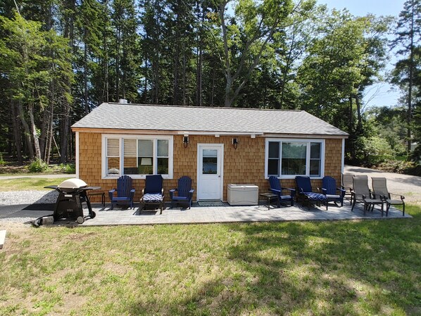 A small wooden cabin with a porch, several Adirondack chairs, and a grill.