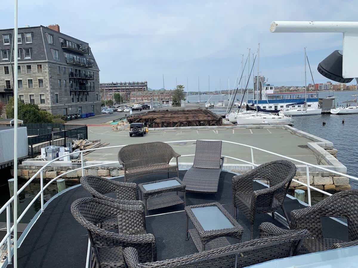 Une terrasse de bateau aménagée avec des meubles en osier, une vue sur le port de plaisance et un bâtiment en arrière-plan.