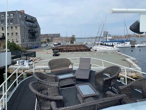 Cubierta de barco con mobiliario de mimbre, vista al puerto deportivo y un edificio al fondo.